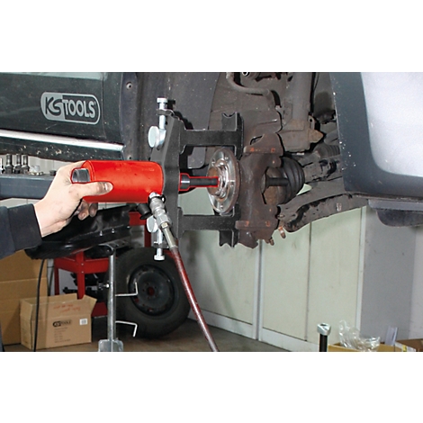 Ein Mechaniker verwendet ein rotes Werkzeug, um an einem Fahrzeug zu arbeiten. Der Fokus liegt auf der Radaufhängung und den Werkzeugen.